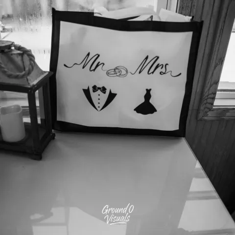 Black and white image of a 'Mr. & Mrs.' gift bag on a table with a candle lantern.