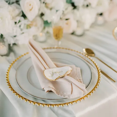 Elegant table setting with gold-rimmed plate, pink napkin, and floral centerpiece.