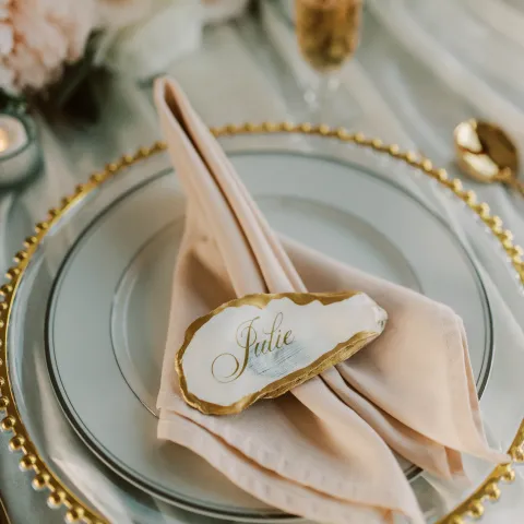 Elegant table setting with gold-rimmed plate, pink napkin, and name card on oyster shell.