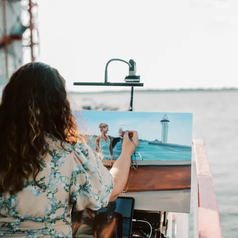 Person painting on a canvas by the water, with a dock visible in the background.