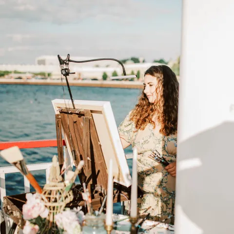 Woman painting on a boat with flowers and candlesticks nearby.