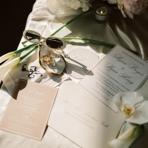 Wedding invitation set with sunglasses, gold rings, and flowers on a table.