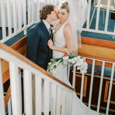 Bride and groom kissing on staircase, holding white flowers.