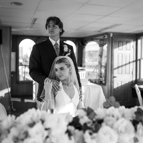 Bride and groom in formal attire on a boat with flowers in the foreground.