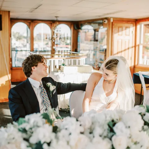 Bride and groom smiling at each other in a decorated room with flowers.