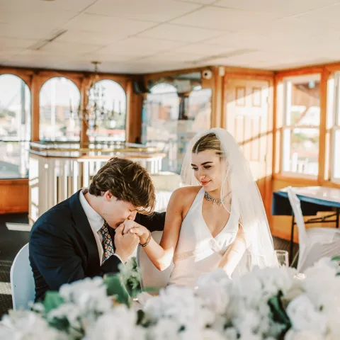 Bride and groom at a table, groom kissing bride's hand, indoors, with floral decorations.