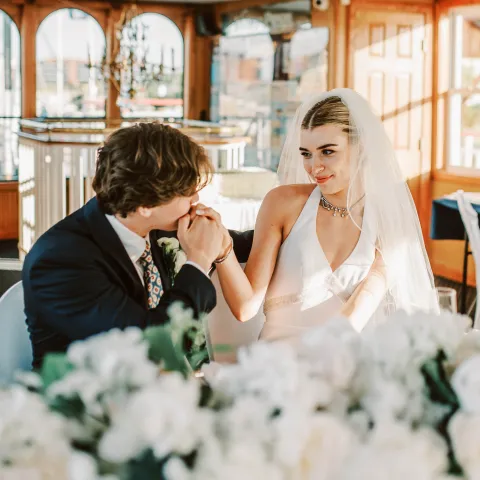 Bride and groom at a table in a warmly lit room, groom kisses bride's hand, flowers in foreground.