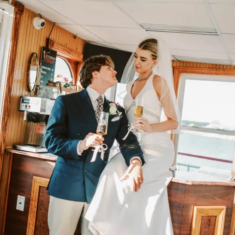 Bride and groom toasting with champagne, bride sitting on counter, indoors.