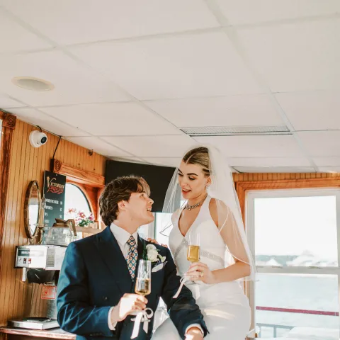 Bride and groom in wedding attire holding champagne glasses on a boat.