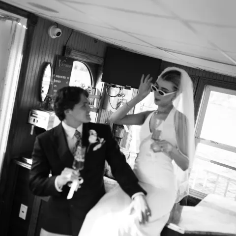 Bride and groom in boat cabin, bride in sunglasses holding a drink.