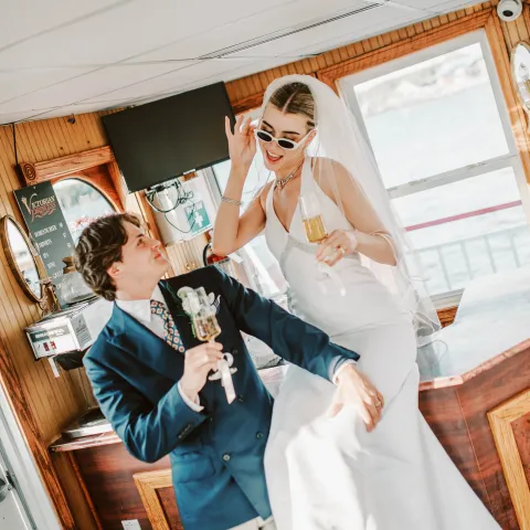 Bride in sunglasses and groom toast with champagne on a boat.