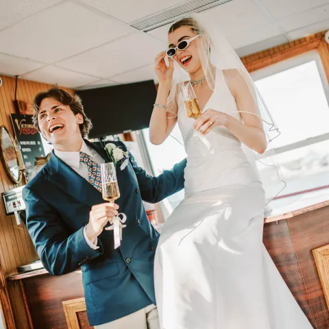 Bride and groom laughing, holding champagne, bride in sunglasses.