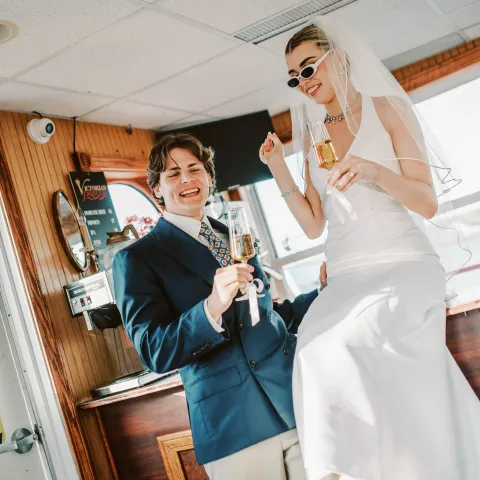 Couple in formal attire celebrating with drinks, woman in white gown and veil, indoors.
