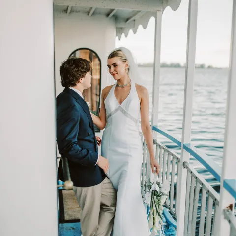 Bride and groom stand on a boat deck overlooking the water.