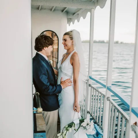 Couple in wedding attire smiling on a boat deck with water in the background.