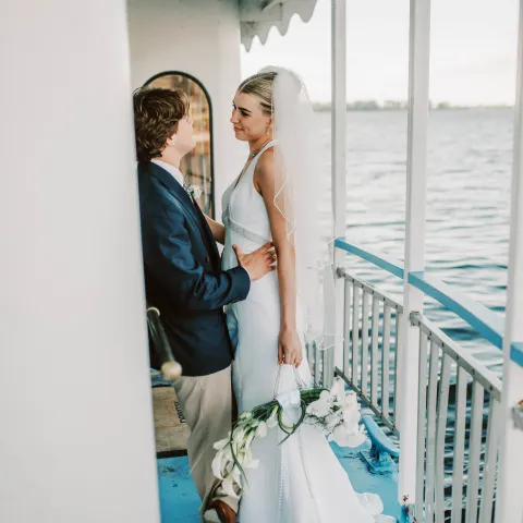 Bride and groom standing on a boat, bride holding flowers, with water view.