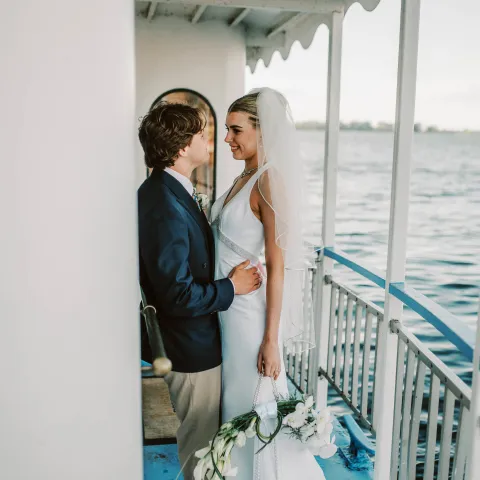 Bride and groom standing on a boat deck, embracing with a water background.