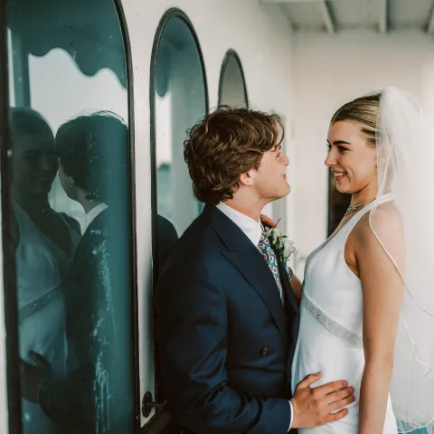 Bride and groom standing close, smiling at each other, with reflections in arched windows.