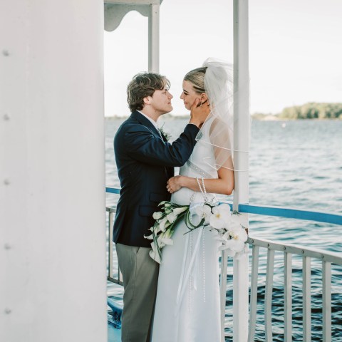 Bride and groom on a boat, standing close with water in the background.