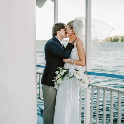 Bride and groom kiss on a boat deck with water in background.