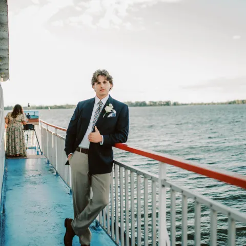 Person in suit leaning on railing of a boat with water and flag in background.