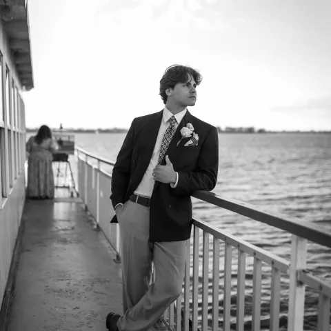 Man in suit leans on boat railing, water in background, American flag flutters above.