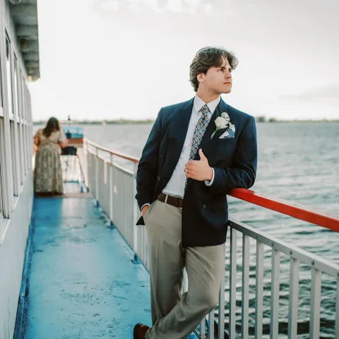 Man in suit leans on railing of a boat with an American flag, ocean in the background.