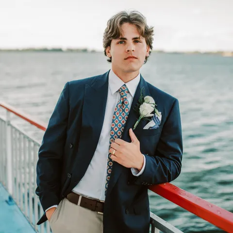 Man in suit with floral boutonniere leaning on boat railing by water.
