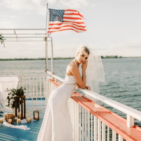 Bride posing on a boat deck with an American flag and water in the background.