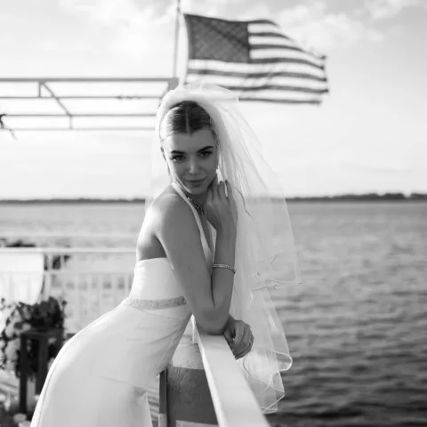 Bride in veil leaning on railing with American flag, near water.