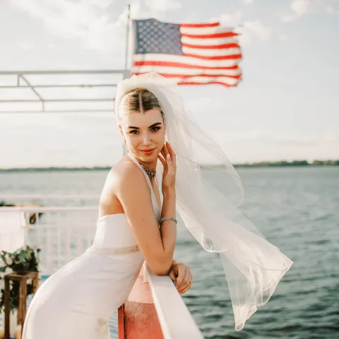 Bride with veil on a boat, American flag in background, water view.