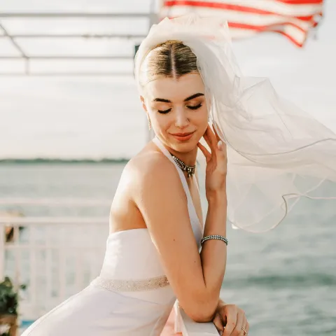 Bride in white dress poses on boat with American flag in background.