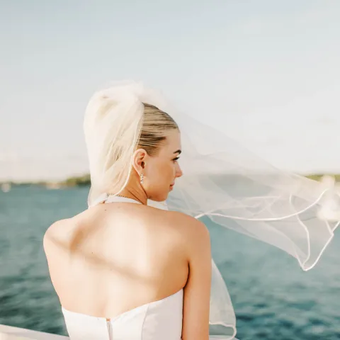 Bride in white dress and veil facing water on sunny day.