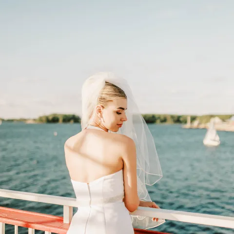 Bride in white dress with veil, standing on a boat railing overlooking a lake.