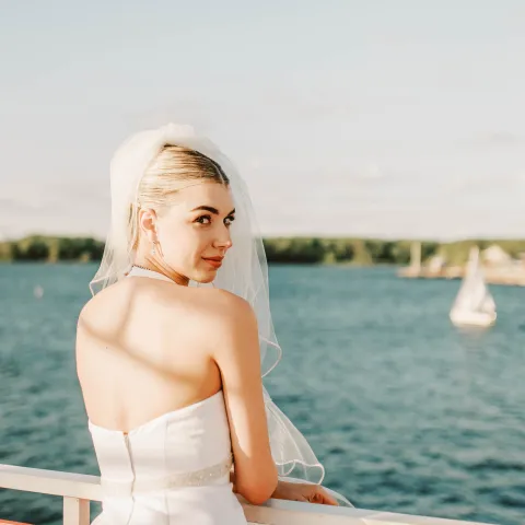 Bride in white dress on balcony overlooking water with sailboat.