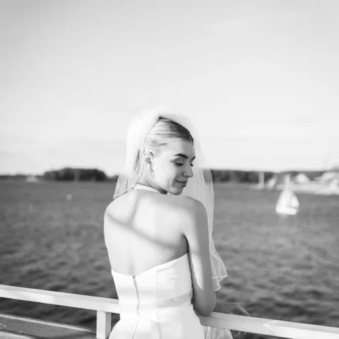 Bride in white gown with veil by water, smiling over shoulder.