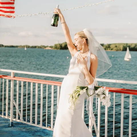 Bride in white gown holding wine bottle on a boat, with lake and American flag in background.