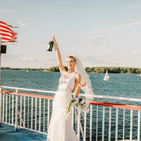 Bride in white dress raising champagne on a boat deck.