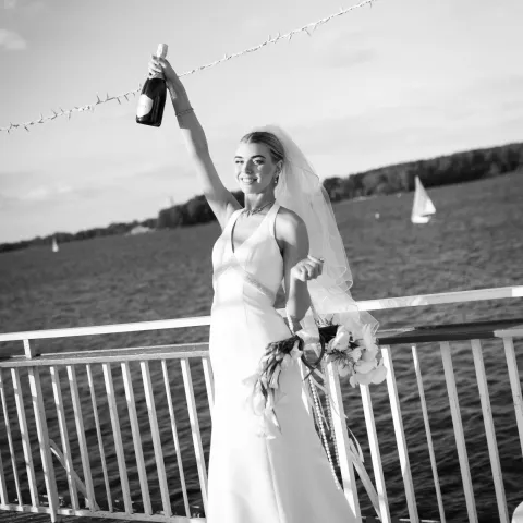 Bride in white dress holding champagne bottle, celebrating on a lakeside pier.