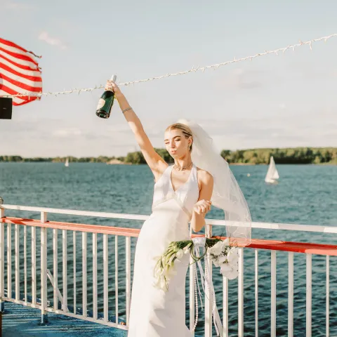 Bride on boat deck celebrating with champagne near water.