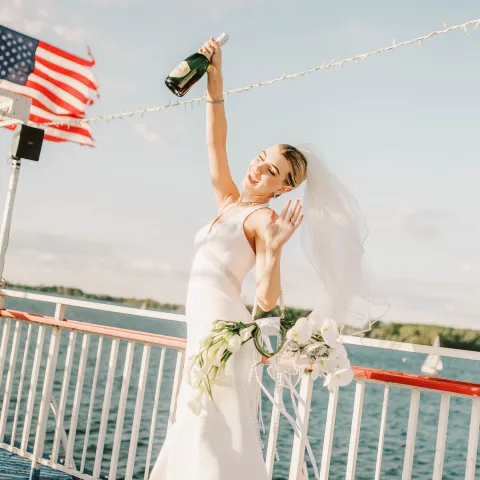 Bride celebrating on a boat with champagne and bouquet, American flag in background.