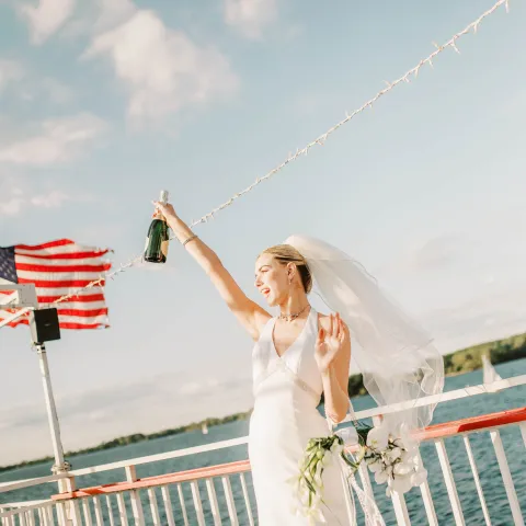 Bride in white dress holding champagne bottle on boat, with a U.S. flag and water in the background.