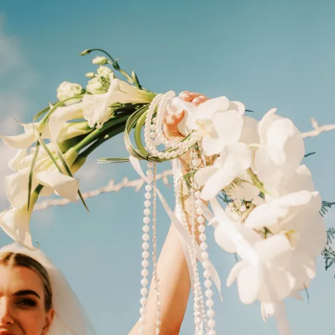Bride holding white flowers and pearls against a blue sky.