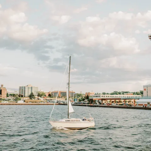 Sailboat on water with cityscape and cloudy sky backdrop.