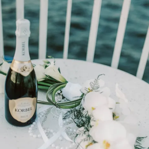 Champagne bottle and flowers on a table by water and railing.