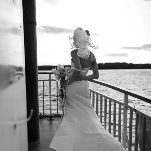 Bride with veil holding bouquet on boat deck by water.