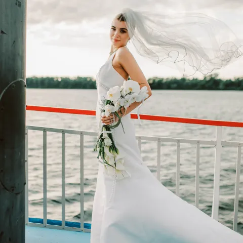 Bride on boat with flowers and veil flowing, water background.