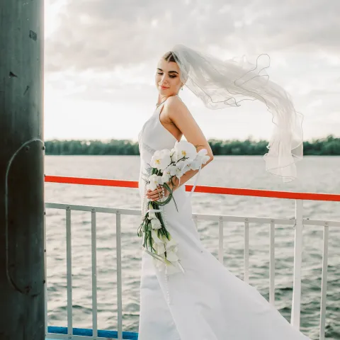 Bride in white dress holding flowers on a boat with veil flowing.