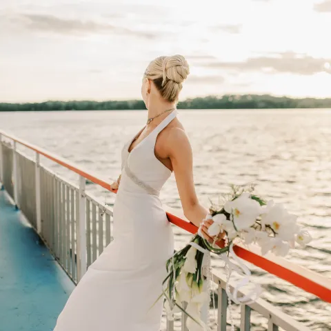 Bride in white dress holding bouquet on boat deck at sunset.