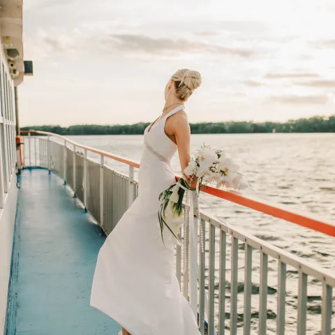Bride in white dress holding bouquet on a boat deck with water view.